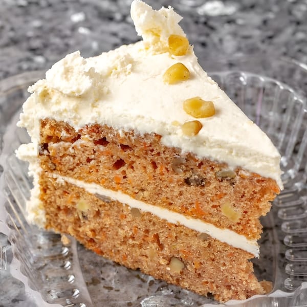 A slice of carrot cake with white frosting and walnuts, in a clear plastic container.