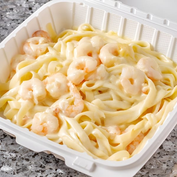 Shrimp Alfredo pasta with creamy white sauce in a white takeout container on a granite surface.